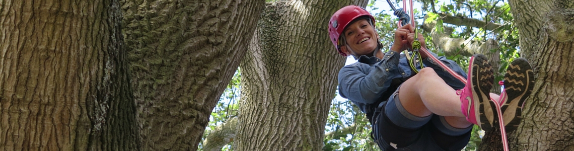 Smiling woman wearing a red helmet and harness, using a rope to climb a large oak tree. She is hanging in between some very large tree branches.