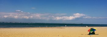 A sandy beach with two people sat under and rainbow coloured sun umbrella