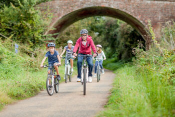 A family group cycling along a cycle path. All are wearing helmets and have just cycled under a brick bridge.