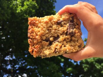 a small hand holds a piece of flapjack with a blue sky and green tree behind