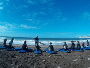 blue sky, sea and beach. people are sat with blue surfboards on the beach. one person is standing in front of them demonstrating. 