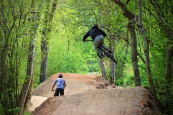 woods with a bumpy path going through them. Two cyclists are riding the trails. 