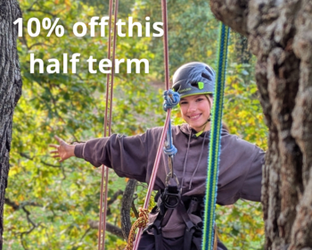 a young woman wearing a helmet and harness has her arms outstretched and is smiling. The background is autumnal leaves and there are ropes hanging in the tree